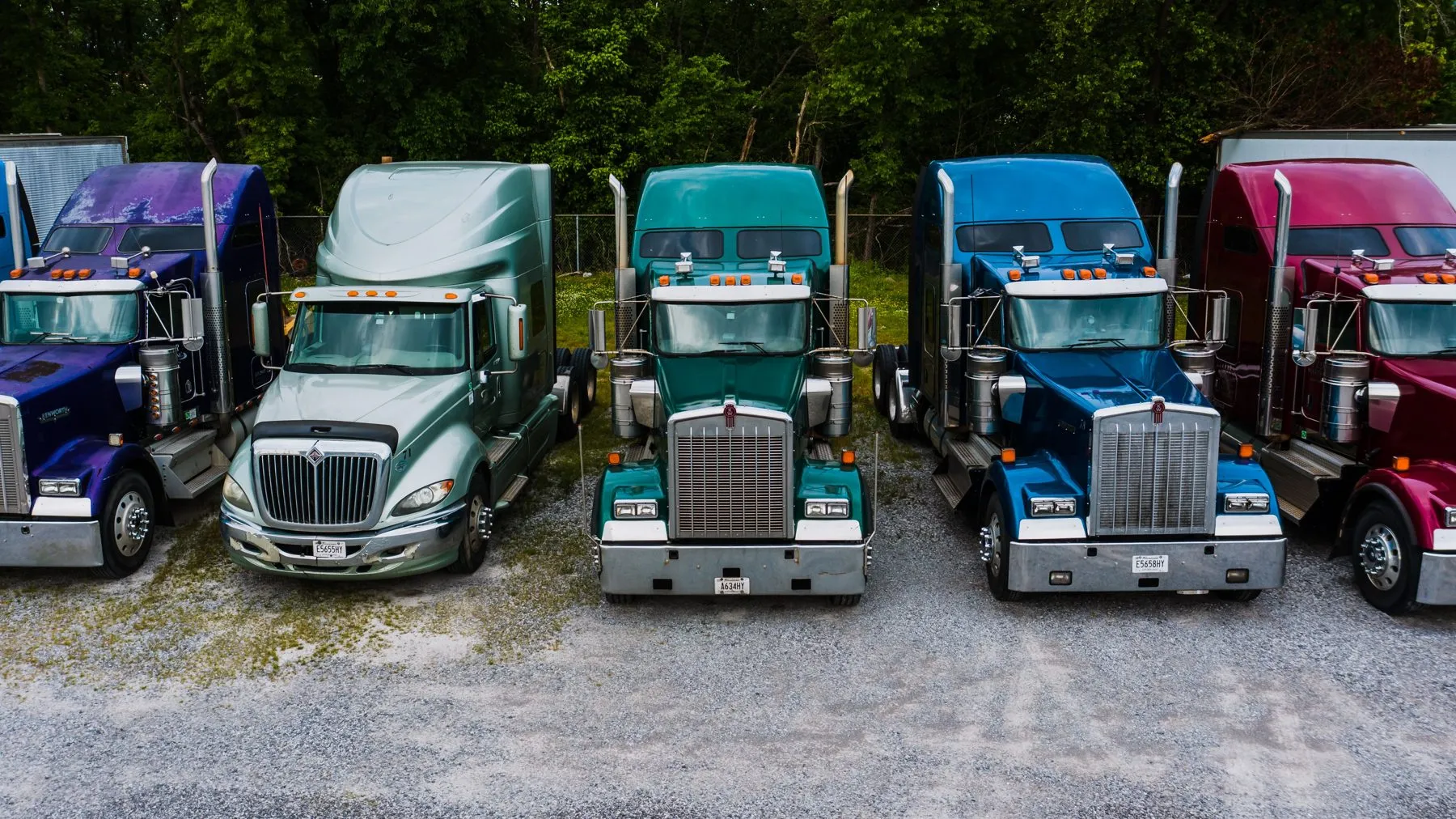 A row of semi tractor cabs parked side by side on a gravel lot in front of trees
