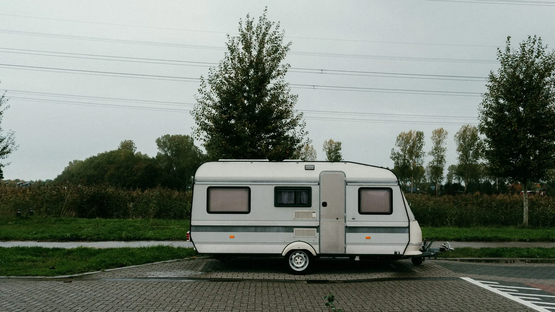 An older white travel trailer parked alone on a paved lot beside a grass verge